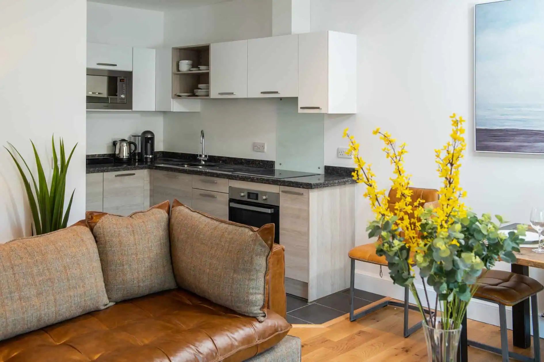 open living space with modern kitchen and brown leather sofa with tweed style cushions and vase of yellow flowers